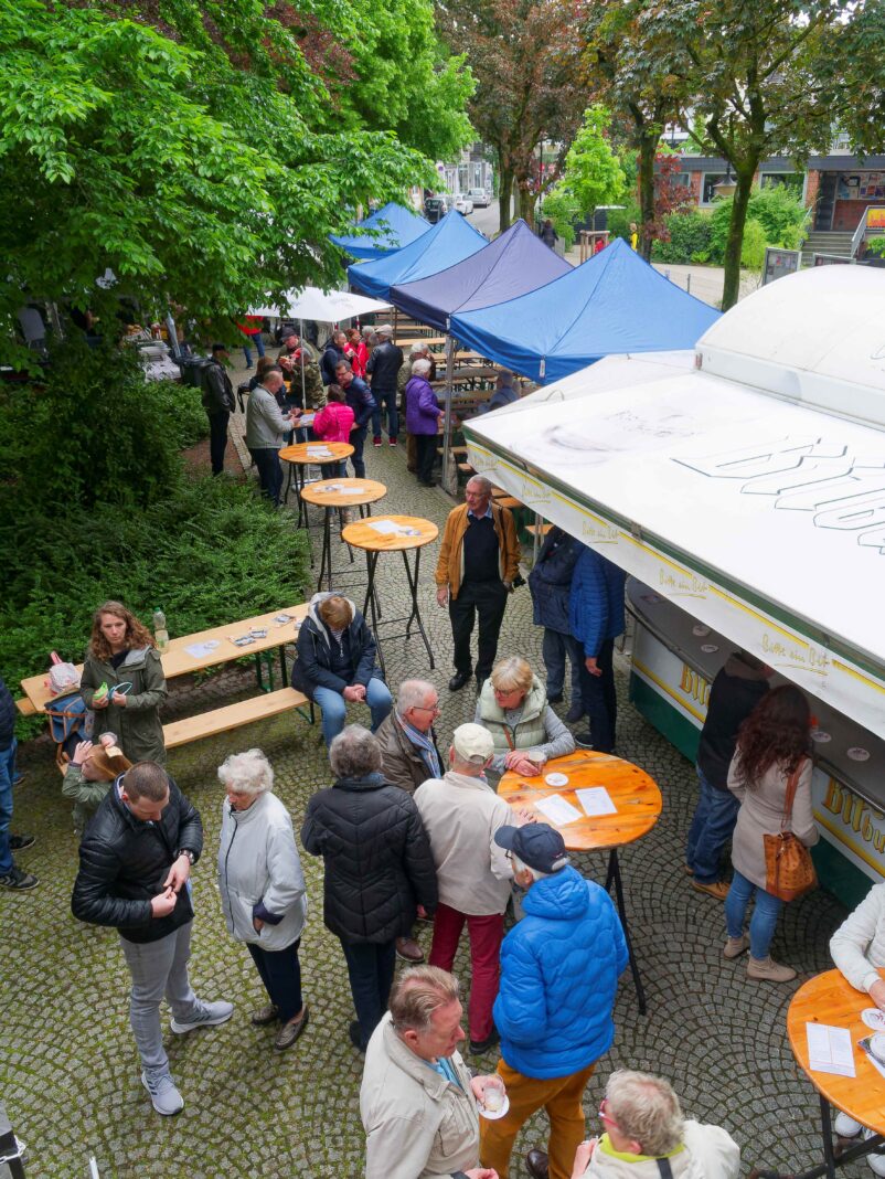 Eine lebhafte Szene auf einem Marktplatz mit vielen Menschen, Stehtischen, blauen Zelten und einem großen Bitburger-Verkaufsstand im Freien.