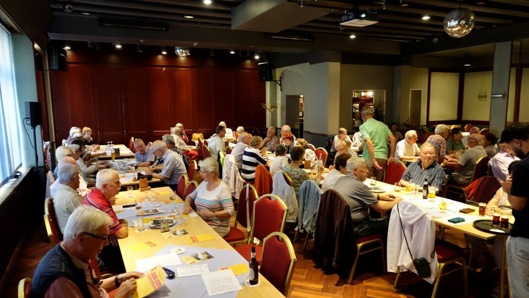 Eine fröhliche Gruppe älterer Menschen sitzt in einem gemütlichen Restaurant an langen Tischen zusammen, unterhält sich und genießt gemeinsam Getränke.