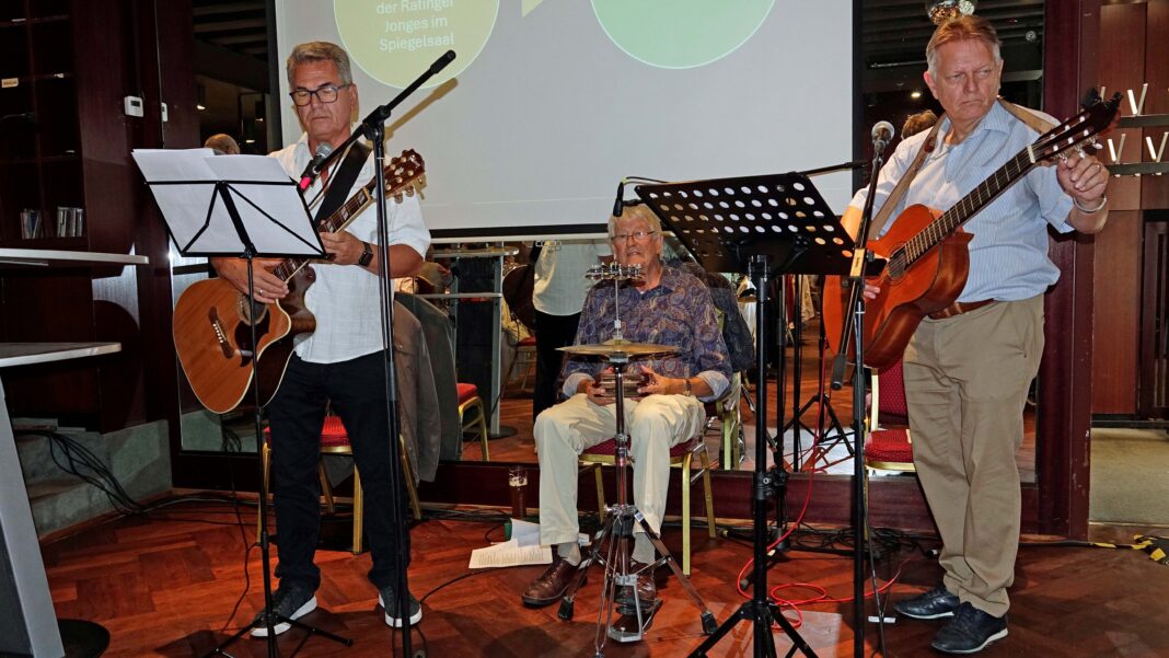 Drei Musiker spielen auf einer Bühne: Zwei Männer stehen mit Akustikgitarren, während ein dritter Mann in der Mitte auf einem kleinen Schlagzeug sitzt.