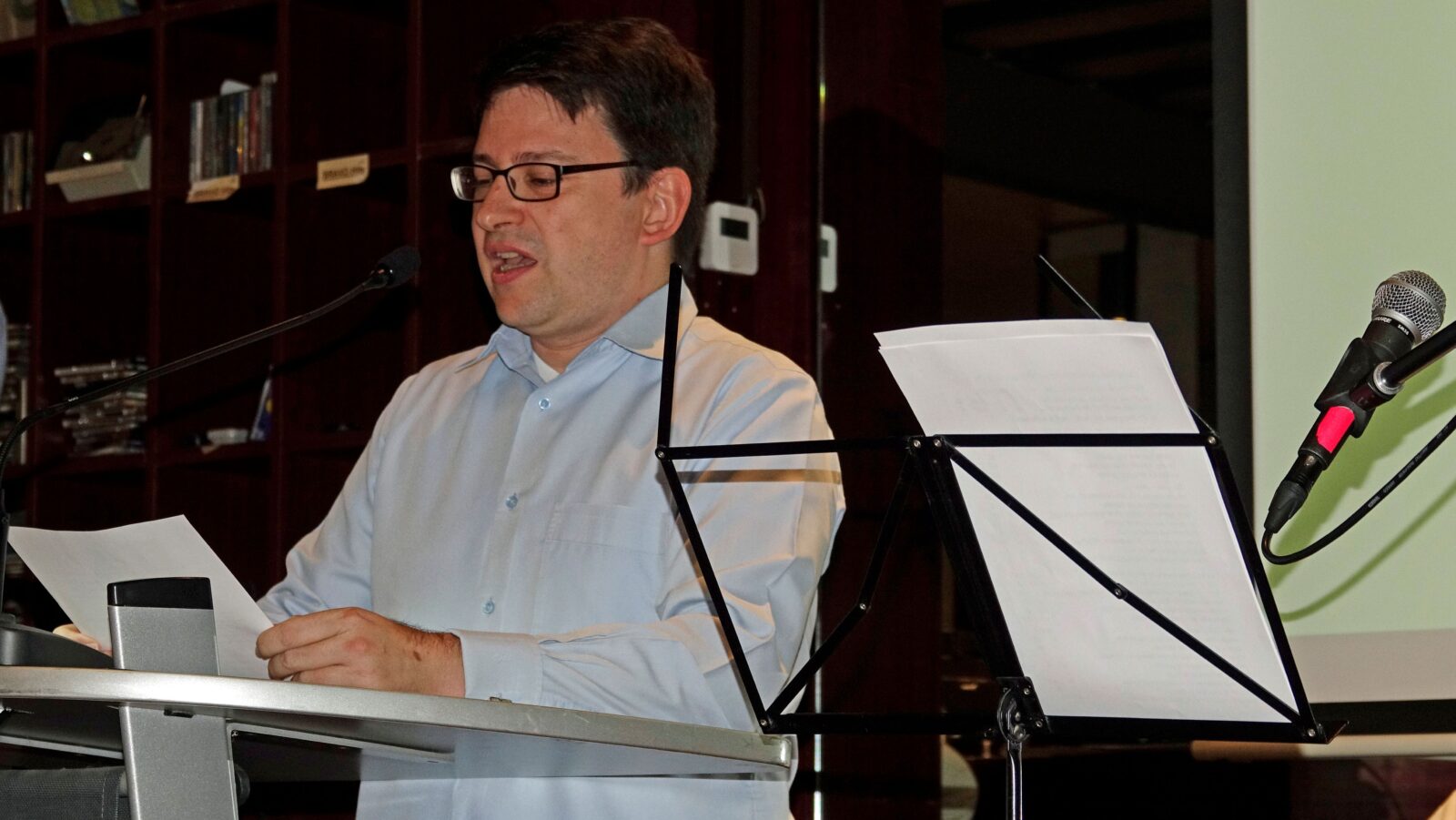 Ein freundlicher Mann mit Brille und hellblauem Hemd hält an einem Rednerpult ein Blatt Papier in der Hand und spricht in ein Mikrofon.
