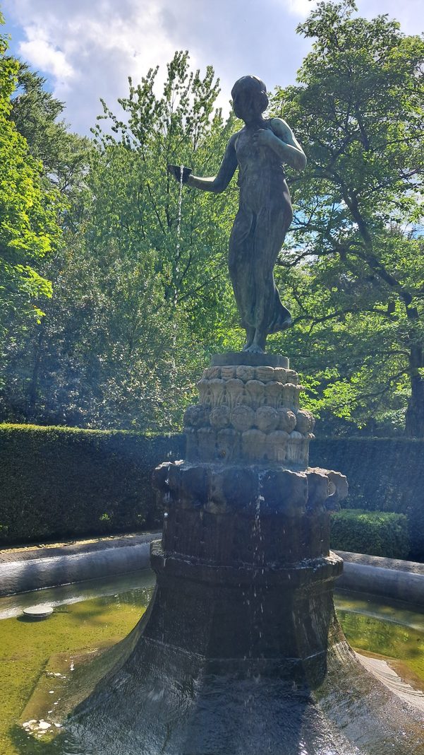 Eine elegante Bronzestatue einer Frau auf einem Brunnen, aus deren Hand Wasser in ein grünes Becken fließt, umgeben von üppigen, sonnigen Bäumen.