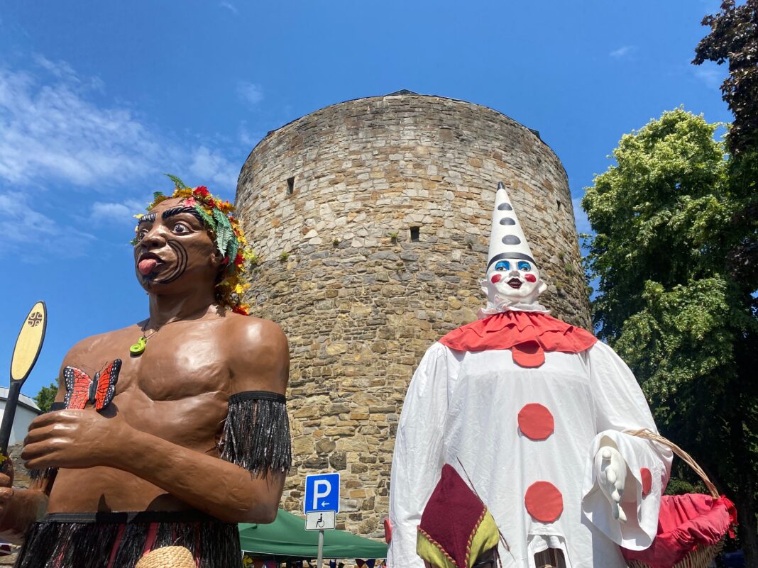Zwei große, bunte Figuren stehen vor einem alten Steinturm unter blauem Himmel. Eine Figur trägt traditionellen Schmuck, die andere ein Clownkostüm.