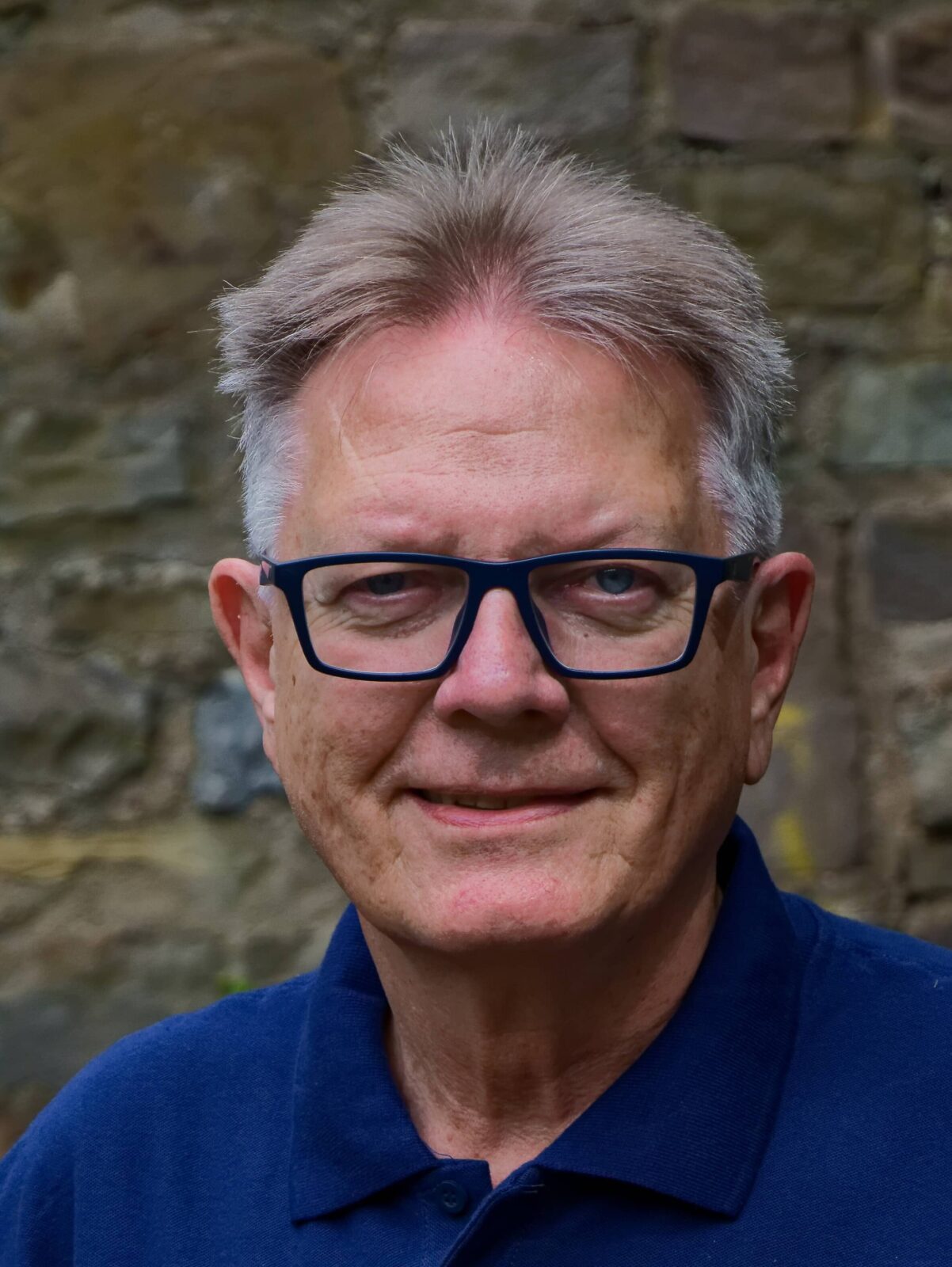 Ein freundlicher Mann mit Brille lächelt vor einer Steinmauer, trägt ein blaues Polo-Shirt mit Logo.