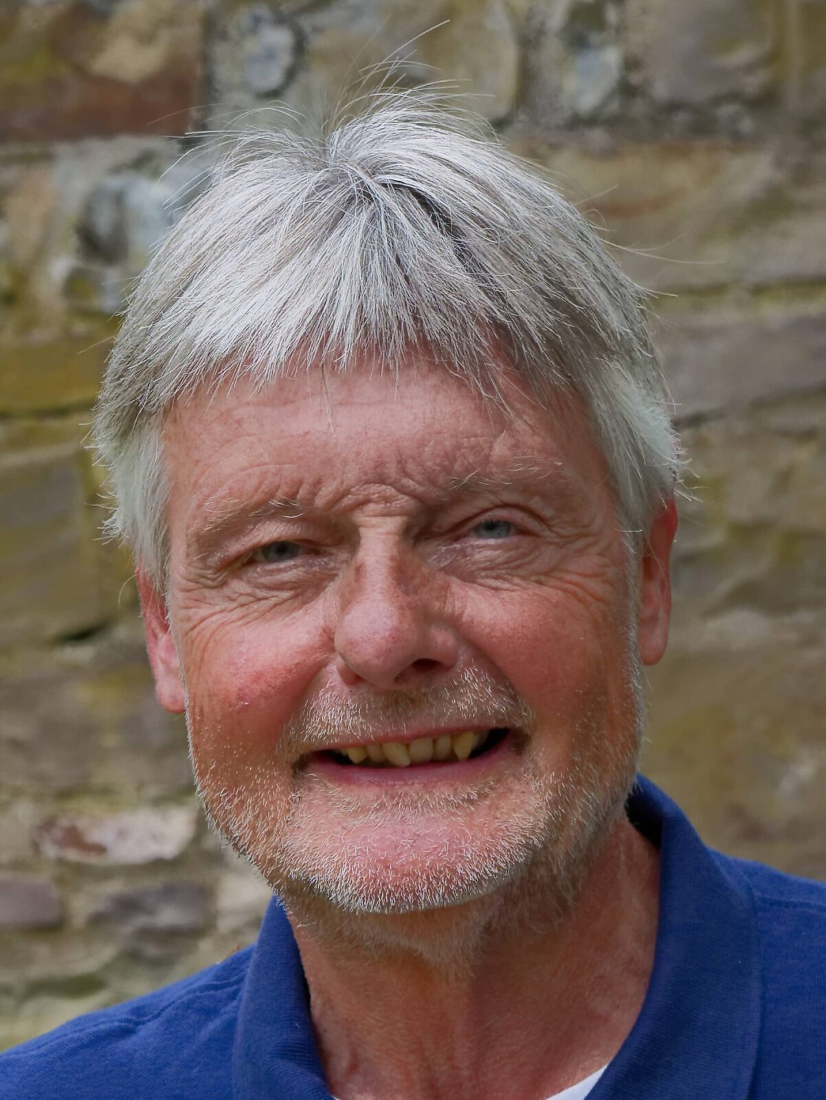 Ein freundlicher Mann mit grauen Haaren lächelt vor einer steinernen Wand. Er trägt ein blaues Polohemd und ein Namensschild.