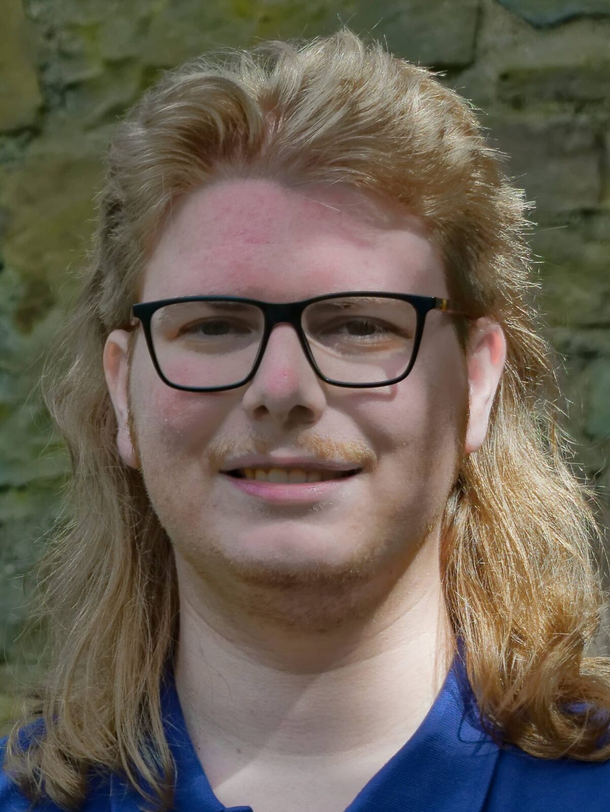 Ein junger Mann mit langen Haaren und Brille lächelt freundlich vor einer Steinmauer.
