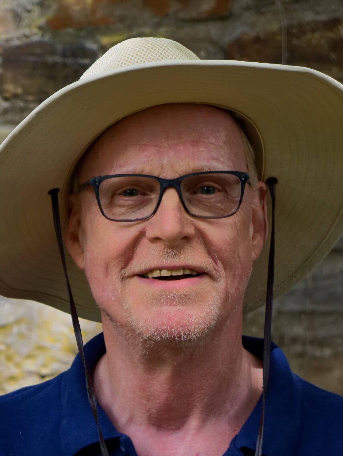 Ein freundlicher Mann mit Hut steht vor einer Steinmauer, lächelt und trägt ein blaues T-Shirt mit einem Logo.