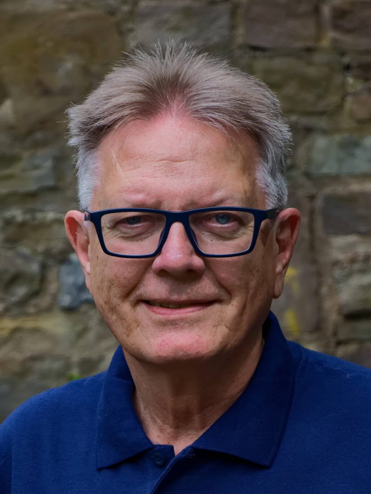 Ein freundlicher Mann mit Brille lächelt vor einer Steinmauer, trägt ein blaues Polo-Shirt mit Logo.