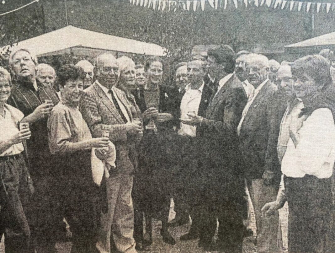 Eine fröhliche Gruppe von Menschen steht bei einer Feier im Freien zusammen, unterhält sich angeregt und hält dabei kleine Gläser in den Händen.