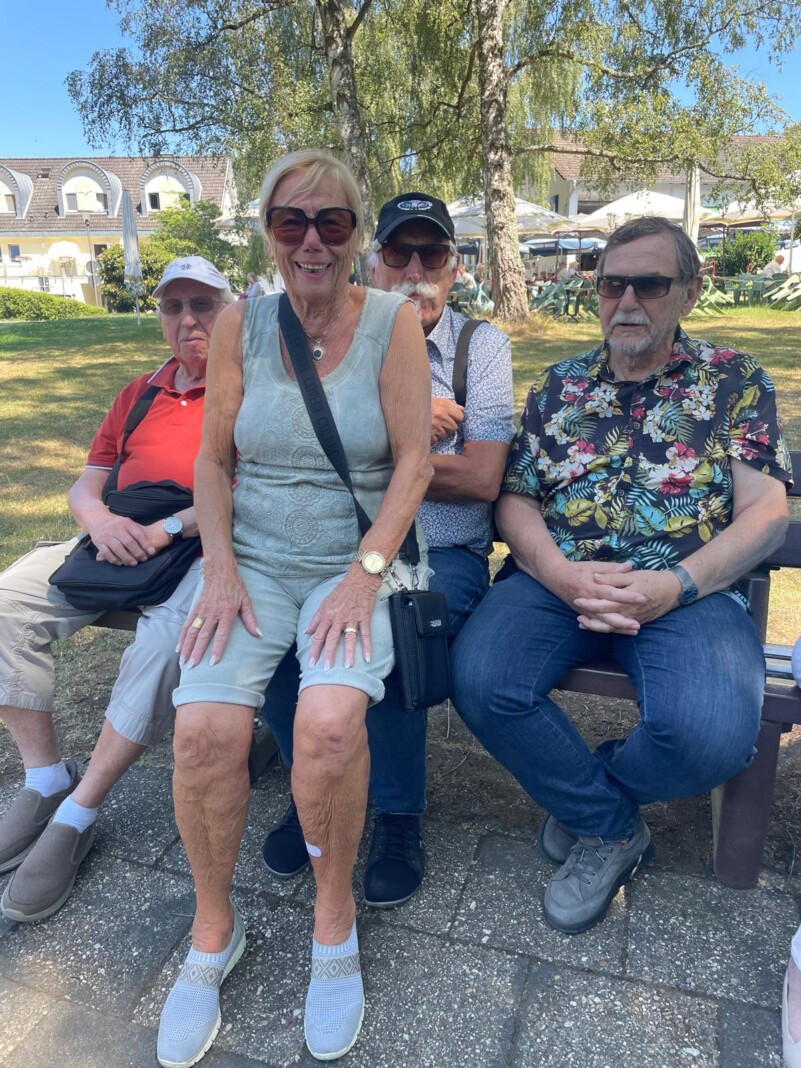 Eine fröhliche Gruppe von vier Senioren sitzt gemeinsam auf einer Parkbank im Freien und genießt bei sonnigem Wetter einen entspannten Tag.