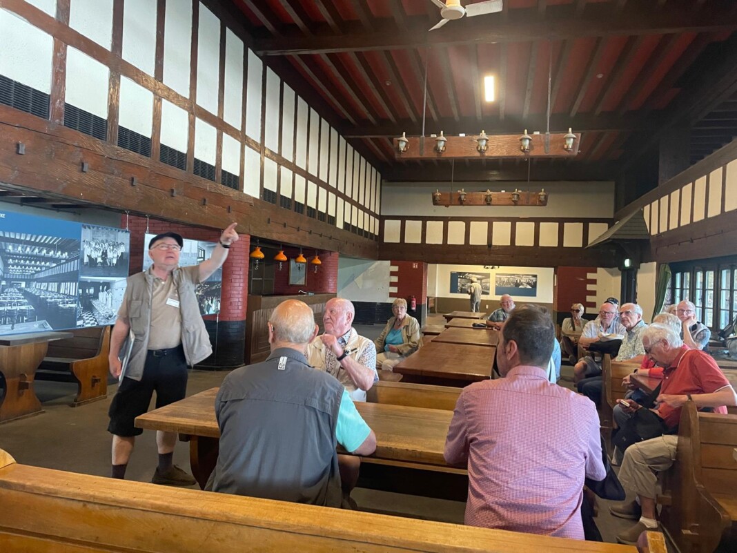 Ein Reiseleiter in Weste zeigt in einem rustikalen Saal mit Fachwerk auf historische Fotos, während eine Gruppe aufmerksam an Holztischen zuhört.