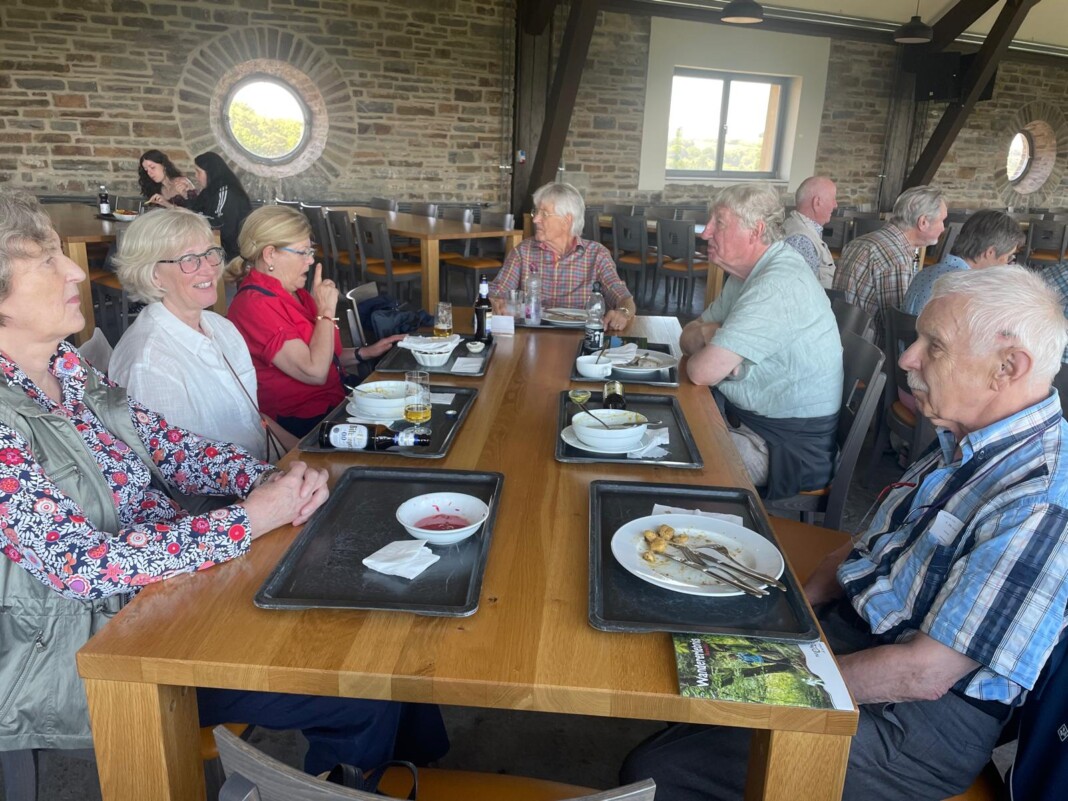 Eine fröhliche Gruppe älterer Menschen sitzt in einem rustikalen Raum mit Steinwänden an einem langen Holztisch beim gemeinsamen Mittagessen.