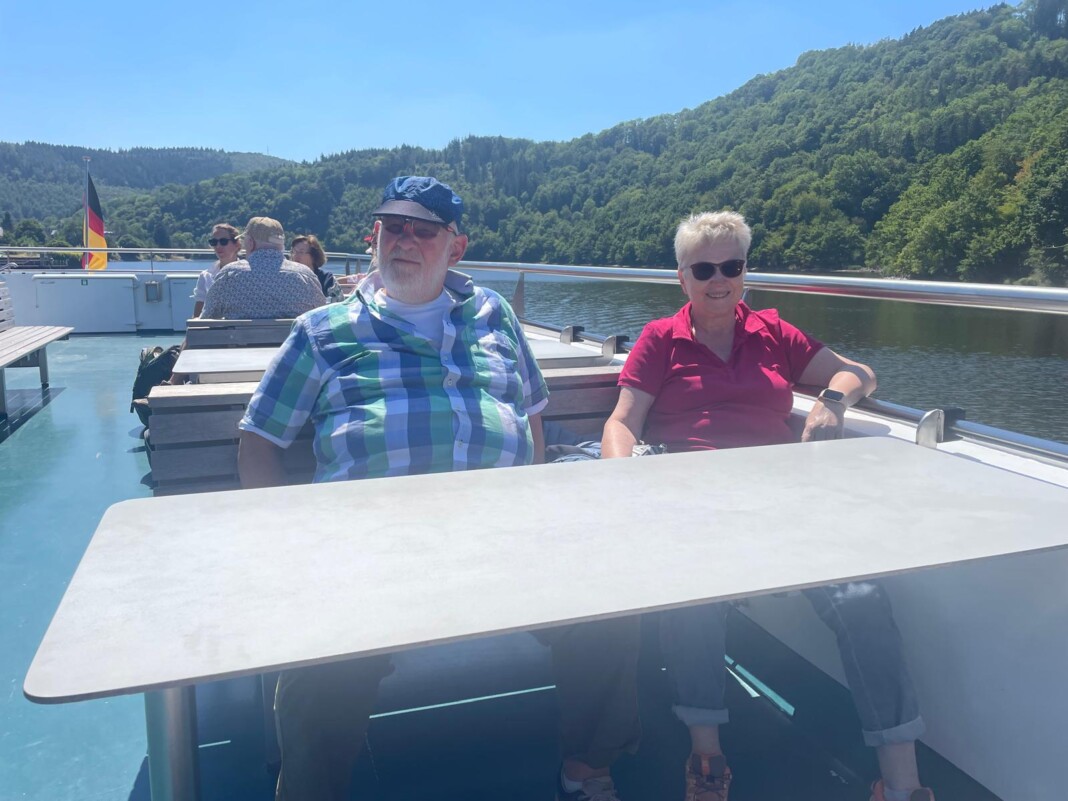Ein freundliches älteres Paar sitzt entspannt an einem Tisch auf dem Deck eines Ausflugsschiffes vor einer sonnigen, bewaldeten Flusslandschaft.