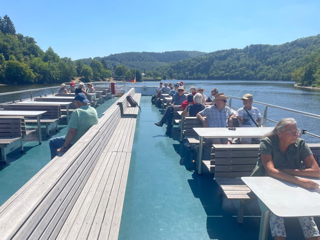 Entspannte Passagiere sitzen an Holztischen auf dem sonnigen Außendeck eines Ausflugsschiffes, das durch eine grüne Hügellandschaft am See fährt.