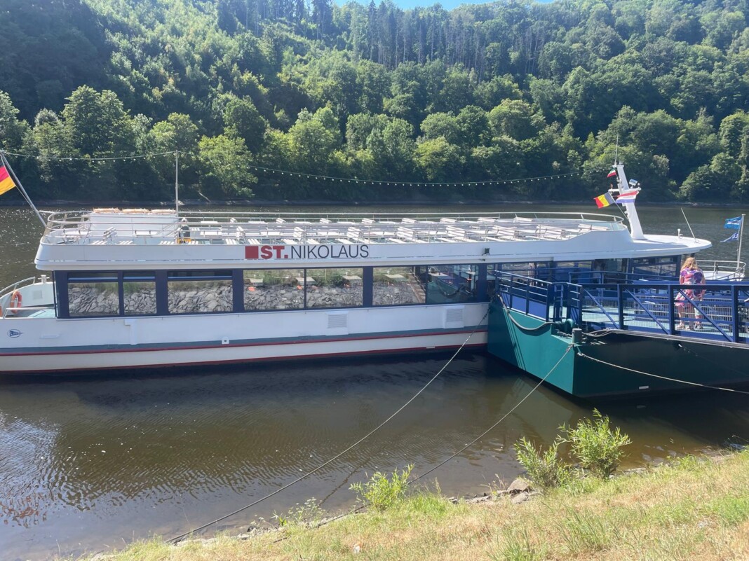 Ein weißes Ausflugsschiff namens St. Nikolaus liegt an einem Bootssteg an einem ruhigen Fluss, umgeben von einem üppig grünen Wald im Hintergrund.