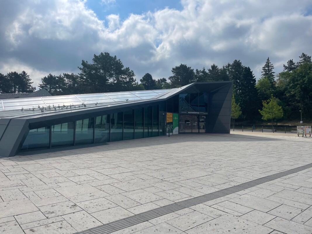 Ein modernes Besucherzentrum mit Glasfassade und schrägem Dach an einem sonnigen Tag, davor ein weitläufiger, gepflasterter Platz mit Bäumen im Hintergrund.