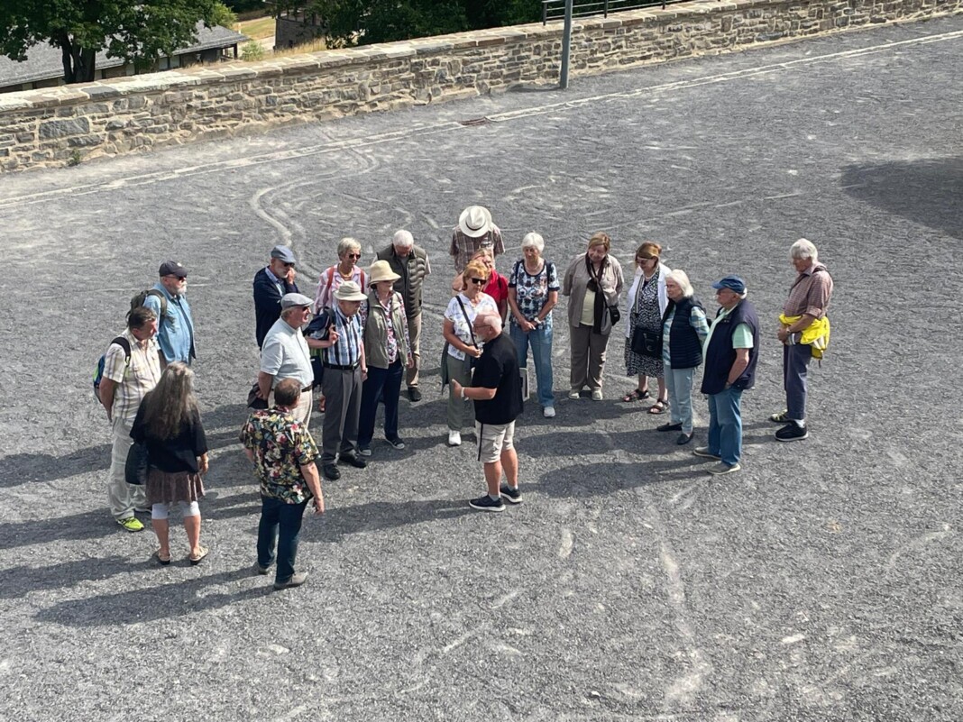 Eine Gruppe von Senioren steht auf einem gepflasterten Platz und hört aufmerksam einem Reiseleiter zu, der ihnen etwas über den Ort erklärt.