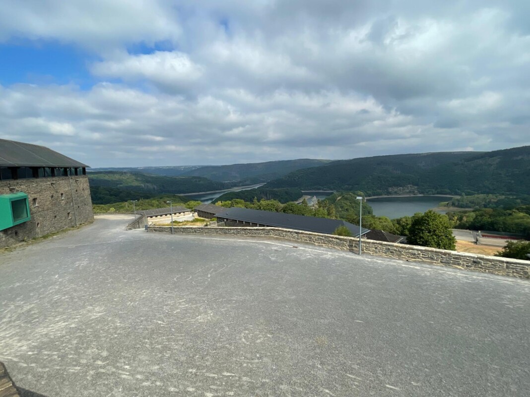 Ein weiter Blick von einer gepflasterten Terrasse auf eine malerische Seenlandschaft, umgeben von grünen Hügeln unter einem bewölkten Sommerhimmel.