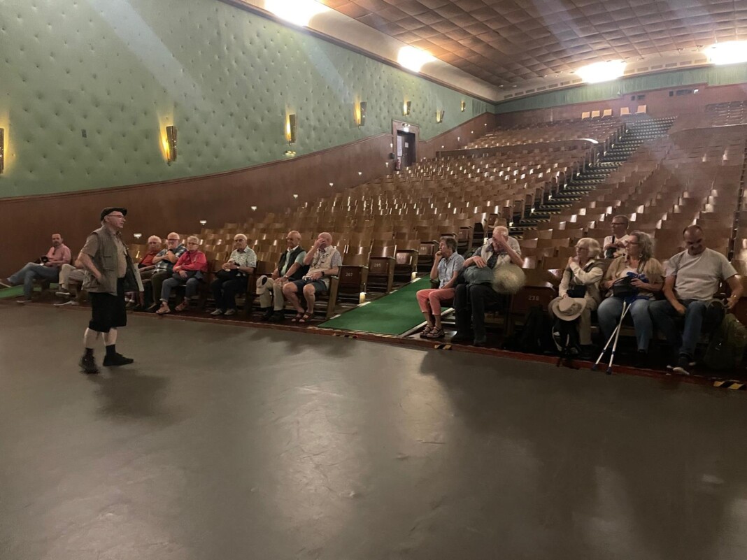 Ein Mann spricht vor einer kleinen Gruppe von Menschen in einem großen, historischen Kinosaal mit vielen leeren Sitzplätzen und grünen Wänden.