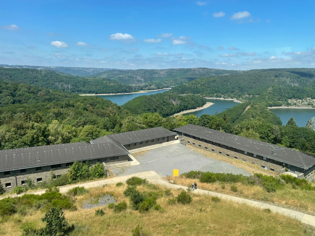 Ein herrlicher Blick von oben auf ein historisches Steingebäude mit Innenhof, umgeben von grünen Wäldern und einem malerischen, geschwungenen Stausee.