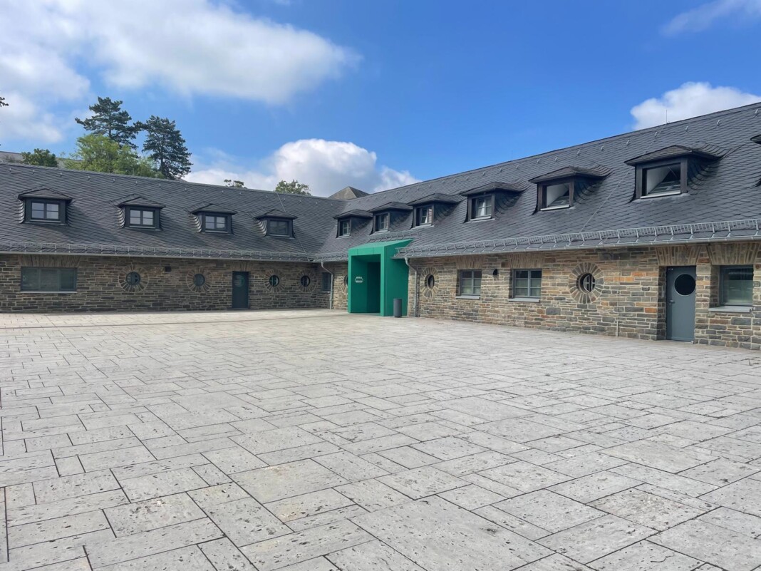 Ein schöner, gepflasterter Innenhof vor einem historischen Gebäude aus Naturstein mit einem auffälligen, modernen grünen Eingangsbereich bei blauem Himmel.