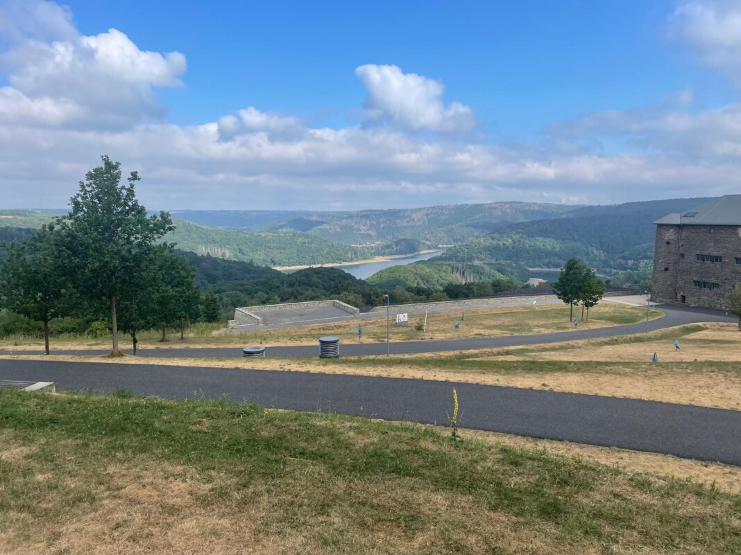 Ein herrlicher Blick auf einen Stausee inmitten grüner Hügel, mit einem gepflasterten Weg und einem steinernen Gebäude an einem sonnigen Sommertag.