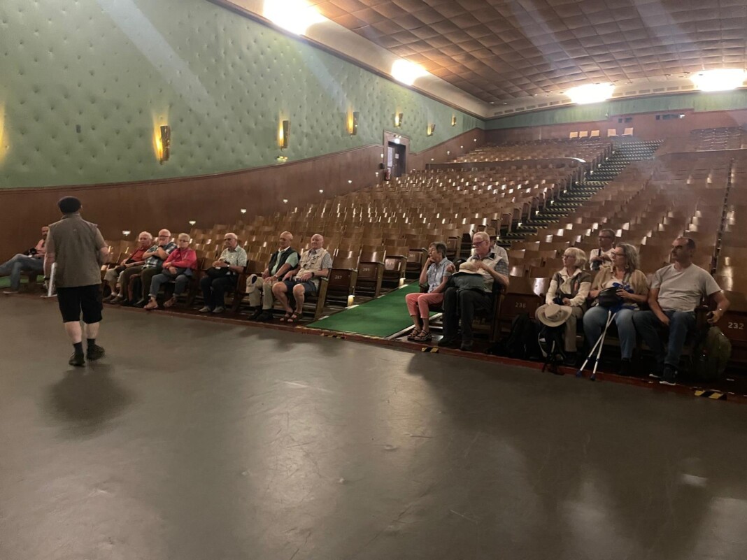 Eine Gruppe von Menschen sitzt in einem großen, historischen Kinosaal und hört aufmerksam einem Sprecher zu, der vor ihnen auf der Bühne steht.