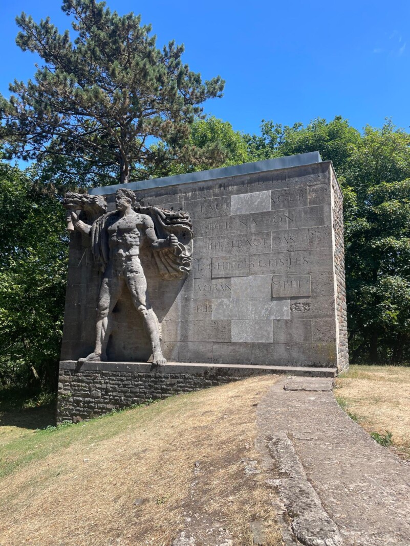 Ein monumentales Steindenkmal mit einer muskulösen Fackelträger-Figur vor Bäumen unter blauem Himmel, mit eingravierten Worten auf der Fassade.
