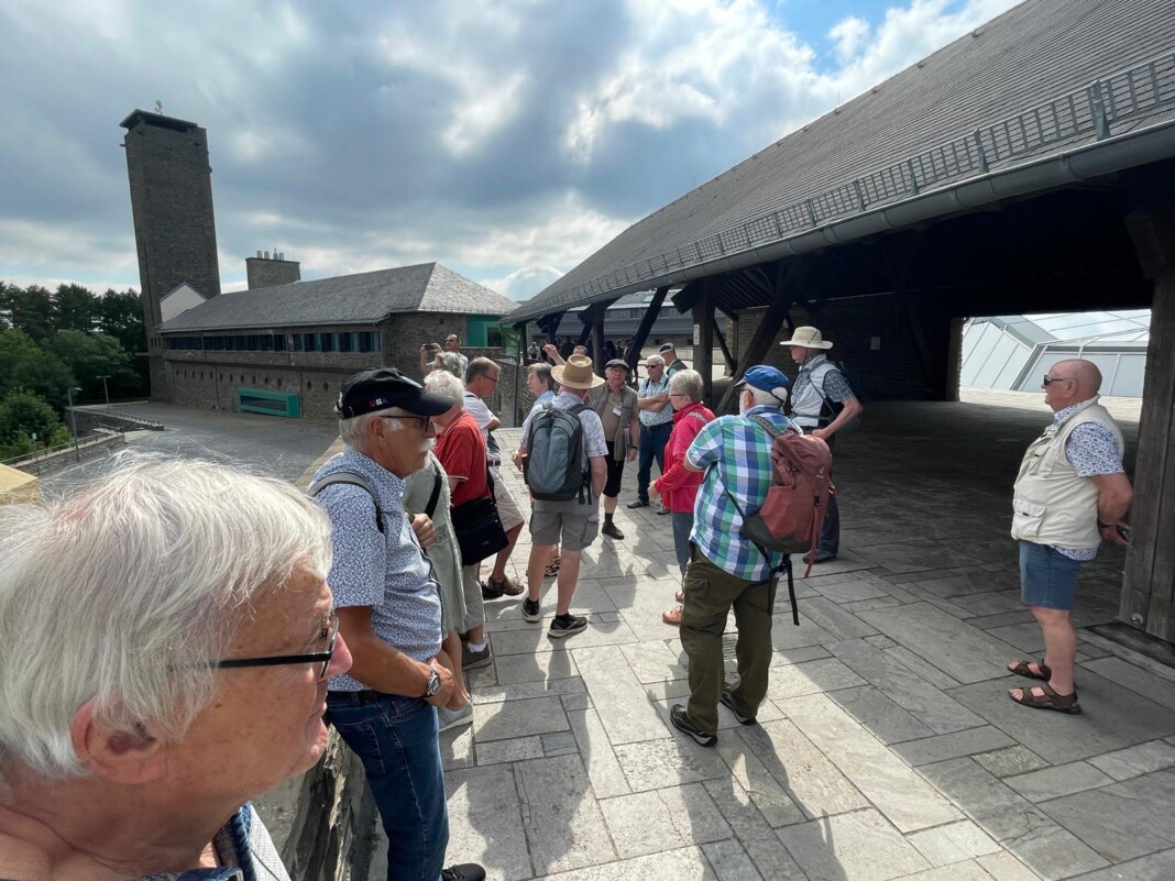 Eine fröhliche Gruppe von Senioren steht an einem sonnigen Tag auf einer gepflasterten Terrasse vor einem historischen Gebäude mit einem hohen Turm.