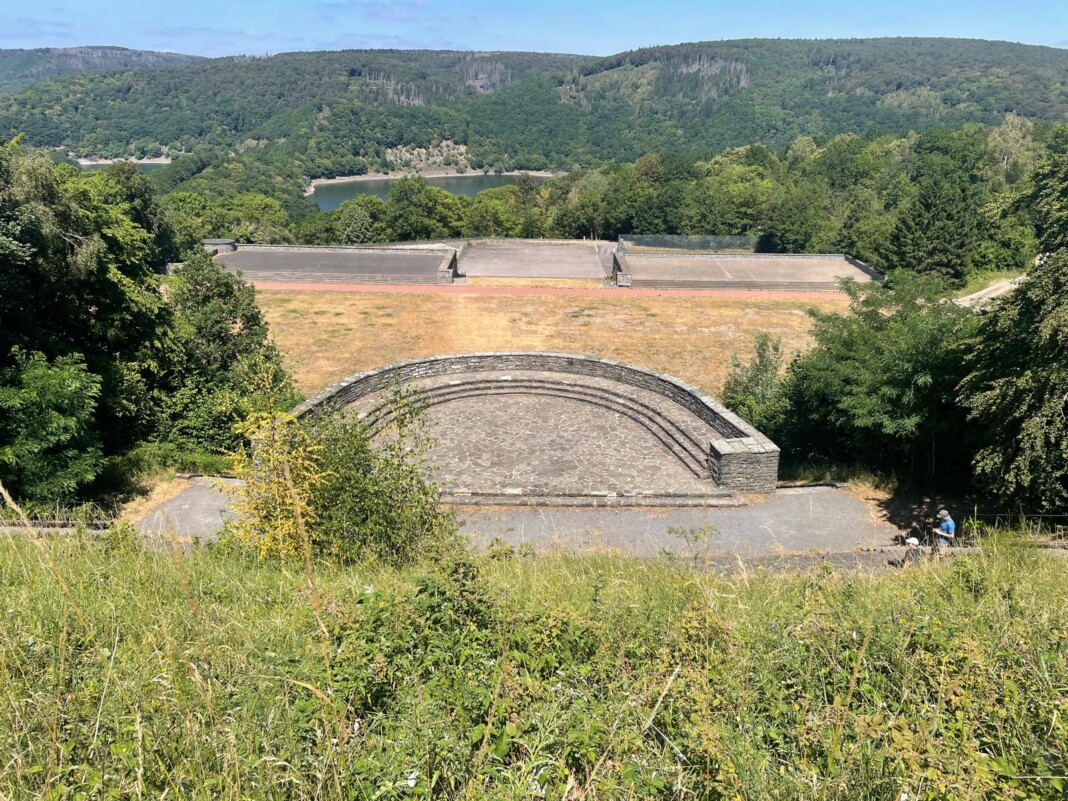 Blick von oben auf eine historische Freilichtbühne aus Stein, umgeben von grünen Wäldern und einem malerischen See in der sonnigen Eifel-Landschaft.
