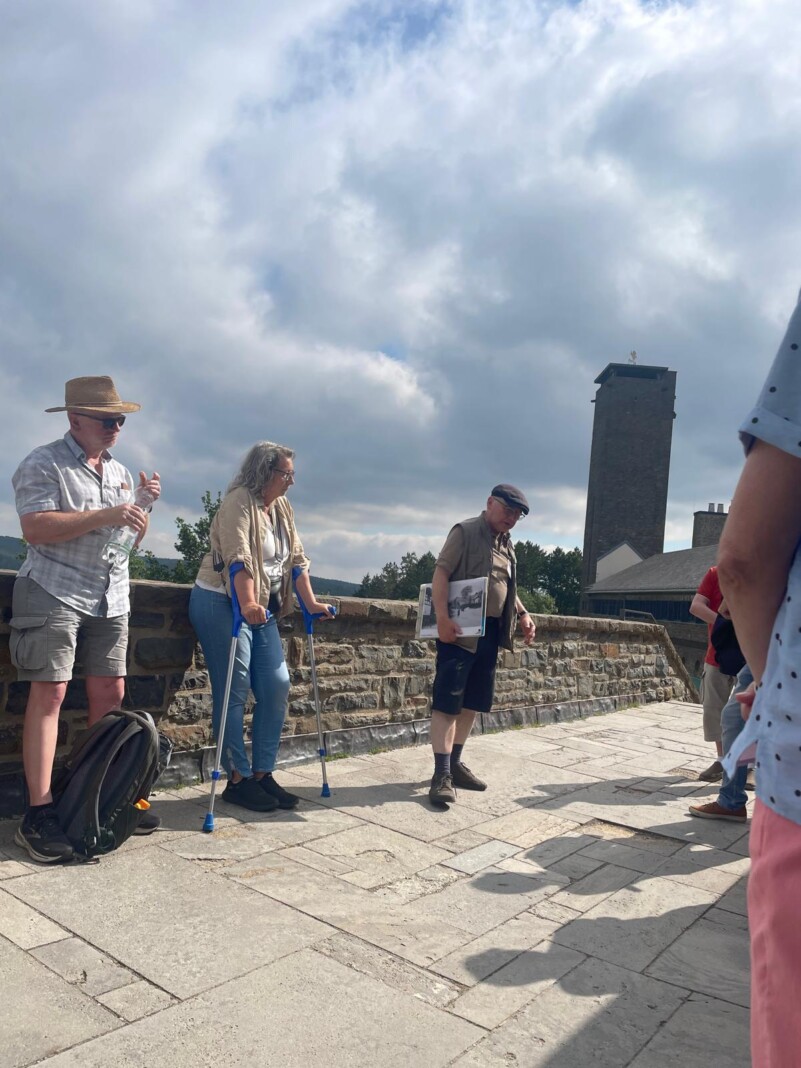 Eine kleine Gruppe von Menschen steht auf einer sonnigen Burgmauer, während ein Mann mit einer Mappe den anderen etwas über die Anlage erzählt.