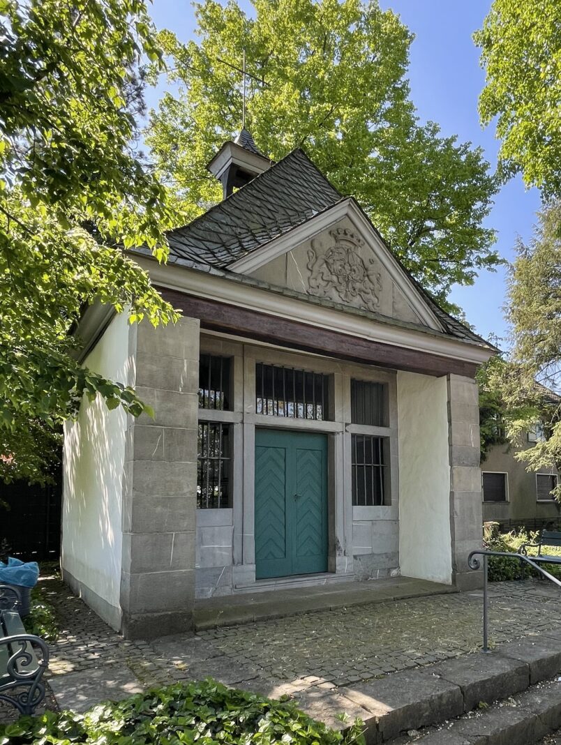 Eine idyllische, kleine Kapelle mit grüner Holztür und einem hübschen Wappen im Giebel, umgeben von sonnigem Grün an einem schönen Sommertag.