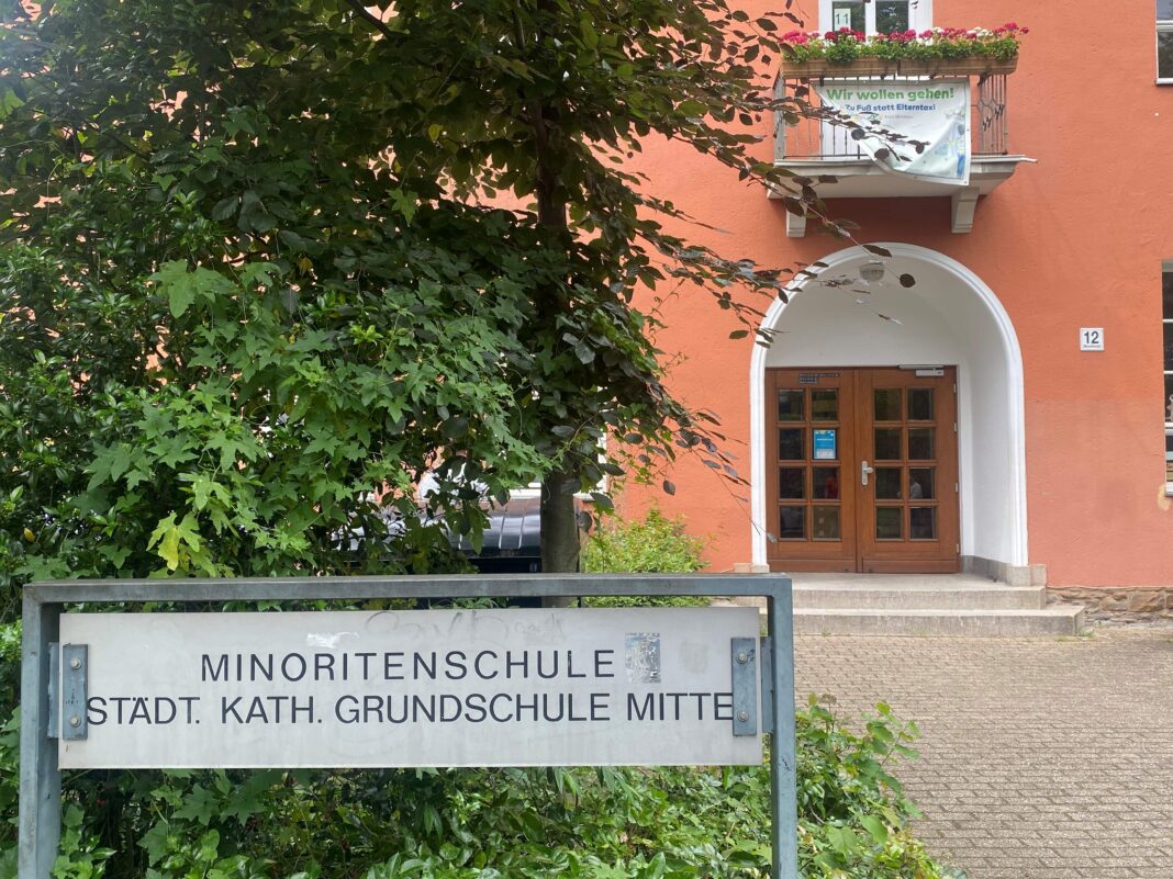 Ein Schild vor der Minoritenschule, einer städtischen katholischen Grundschule, mit einem Balkonbanner, das zum Gehen statt zum Elterntaxi aufruft.