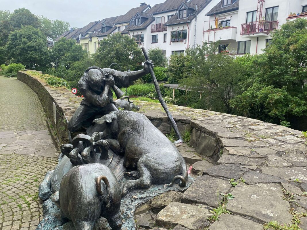 Eine bronzene Statue eines Schweinehirten mit seinen Tieren auf einer Steinmauer vor einer Wohnsiedlung an einem sonnigen Tag in einer Stadt.