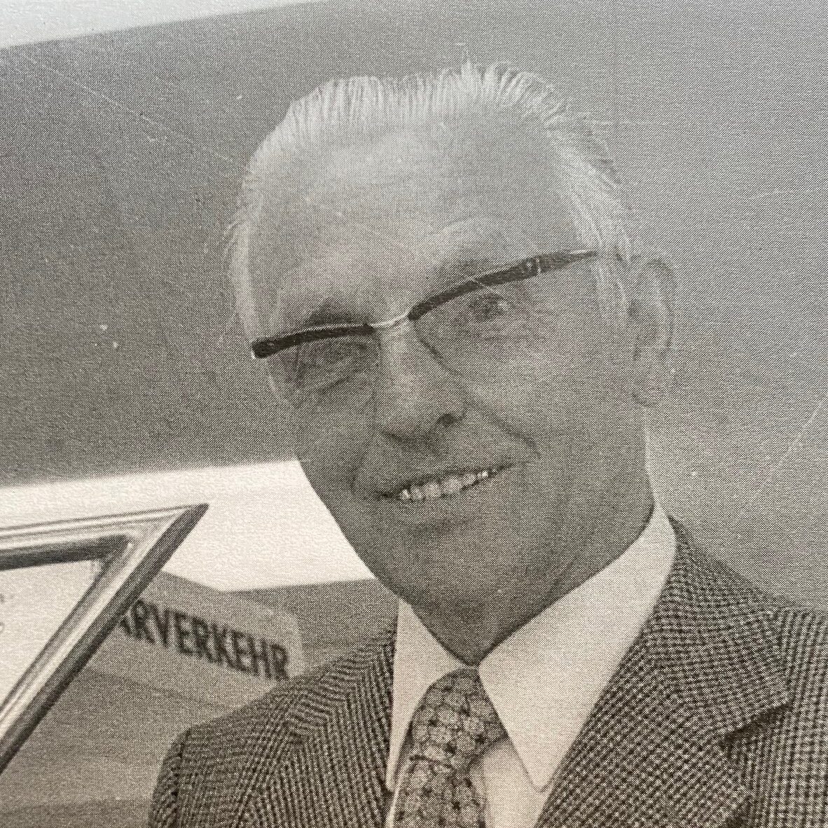 Ein freundlicher älterer Herr mit Brille, Hemd und Krawatte lächelt in die Kamera. Im Hintergrund ist ein Schild mit dem Wort KONTOER zu sehen.