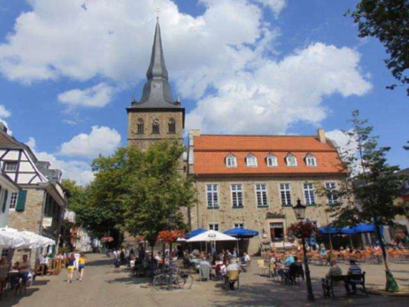 Ein sonniger Marktplatz mit einer historischen Kirche, Fachwerkhäusern und vielen Menschen, die in gemütlichen Straßencafés unter Sonnenschirmen sitzen.