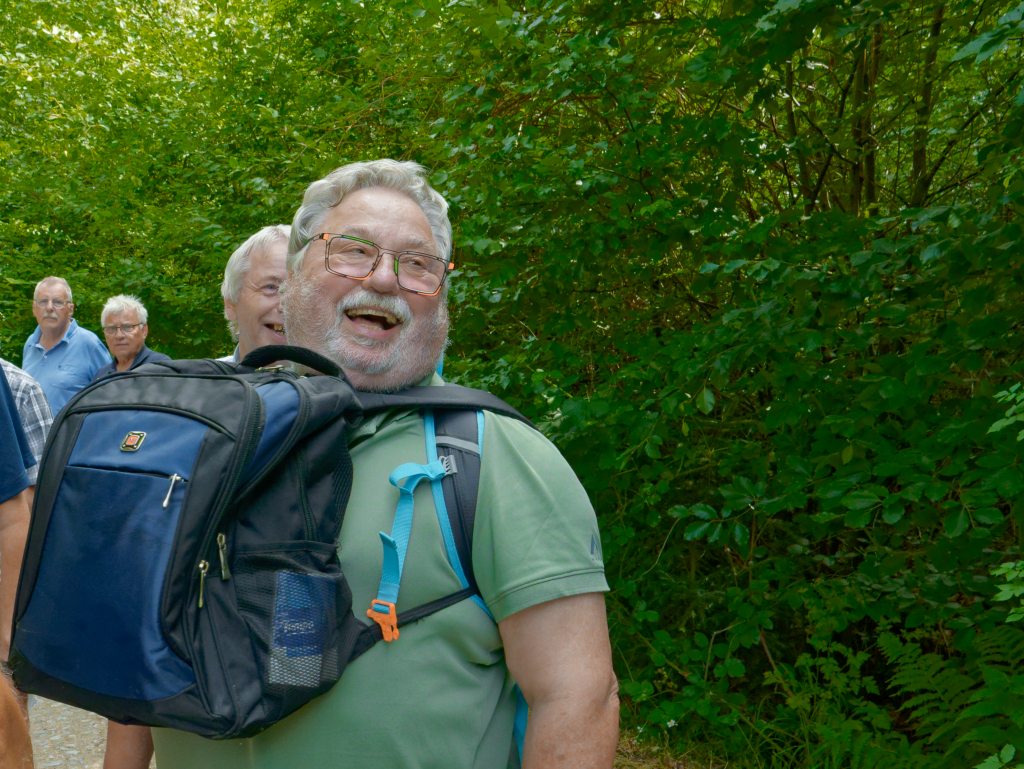 Ein fröhlicher, älterer Mann mit Brille und Rucksack lacht herzlich bei einem entspannten Spaziergang im Grünen mit seinen Freunden im Hintergrund.