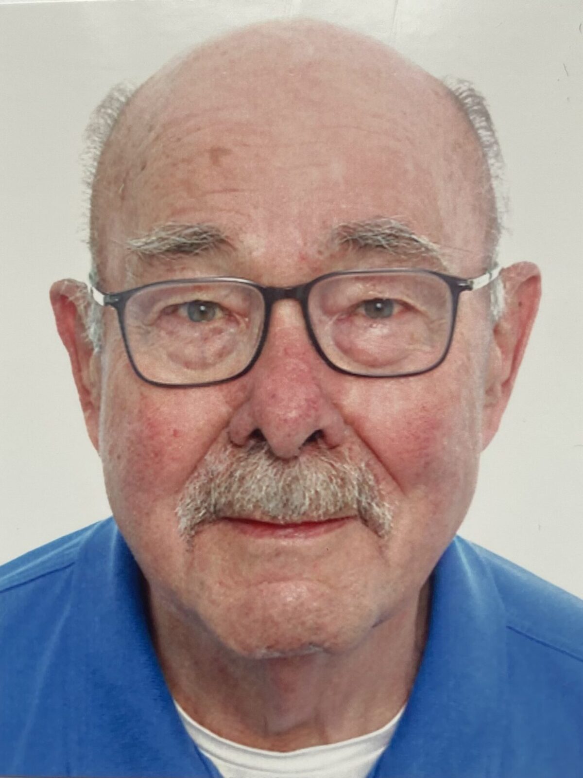 Ein älterer Mann mit Brille und einem freundlichen Lächeln, trägt ein blaues Hemd und hat einen grauen Schnurrbart.